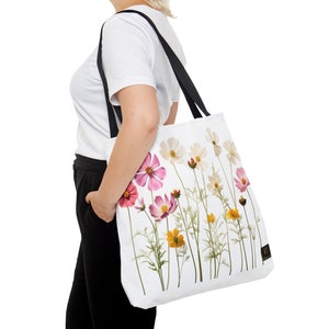 Puede incluir: Un bolso tote blanco con un estampado botánico de varias flores cosmos en tonos rosa, blanco y amarillo. El bolso tiene dos correas negras para el hombro y una pequeña etiqueta de marca rectangular negra en la parte inferior derecha.