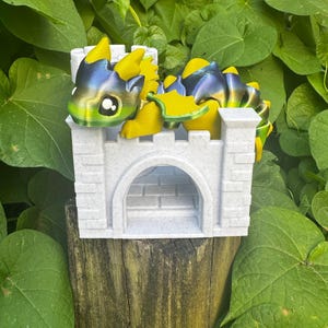 Peut inclure: Un jouet dragon imprimé en 3D avec des écailles jaunes et bleu irisé, niché dans un château blanc. Le château a une porte voûtée et des murs crénelés. Le jouet est placé sur un fond de feuillage vert luxuriant et un poteau en bois vieilli.