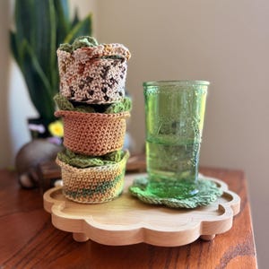 May include: A stack of three crocheted cup sleeves in variegated earth tones of brown, tan, and green, positioned next to a green glass tumbler resting on a matching crocheted coaster, all set upon a light-colored wooden scalloped display tray.