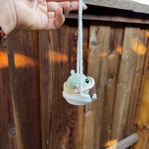 May include: A small green crocheted character, resembling a baby, is sitting in a crocheted swing. The swing is hanging from a white string.