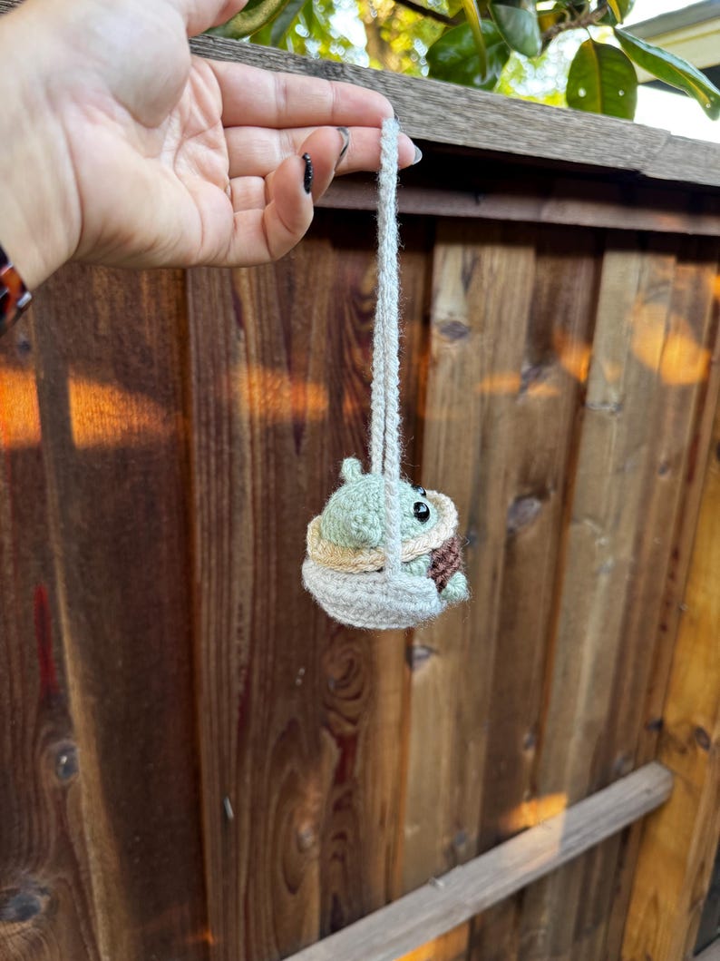 May include: A small green crocheted character, resembling a baby, is sitting in a crocheted swing. The swing is hanging from a white string.