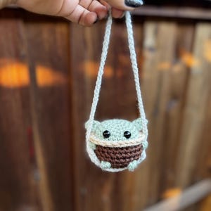 May include: A small crocheted green and brown character with large eyes hanging from a white string. The character is wearing a brown hat and has a small mouth.