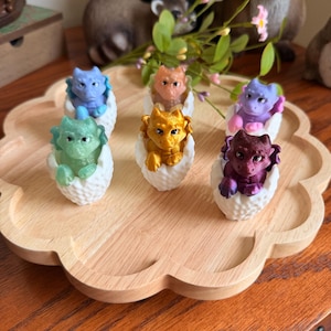 May include: Six small dragon figurines in various colors, including blue, green, orange, gold, purple, and dark red, each emerging from a textured white egg shell. The dragons have visible wings, horns, and detailed facial features, arranged on a wooden tray.