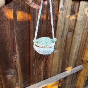 May include: A small crocheted bag with a green character design. The bag has a white strap and is hanging from a hand.