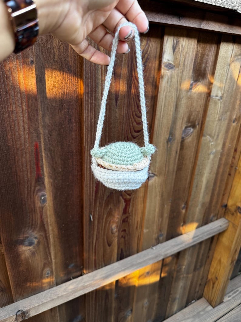 May include: A small crocheted bag with a green character design. The bag has a white strap and is hanging from a hand.