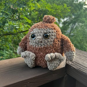 May include: A brown crocheted plush toy with large brown eyes and a small brown pom-pom on its head. The toy is sitting on a wooden surface.
