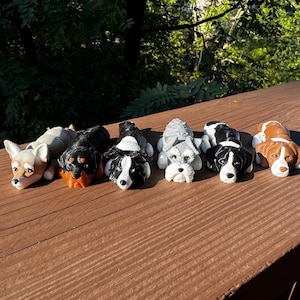 May include: Seven small, realistic-looking dog figurines, each in a different breed and colour, lying down on a wooden surface. The dogs are made of painted resin and have detailed features.