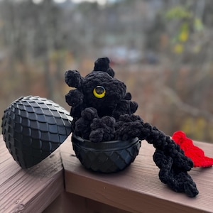Peut inclure: Un jouet en peluche de dragon noir crocheté avec un œil jaune, assis dans un œuf de dragon noir. L'œuf est ouvert et la queue du dragon pend.
