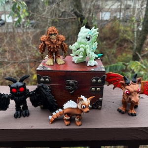 May include: A collection of six small, hand-painted figurines, including a brown yeti, a green monster, a black bat, a brown dragon, a brown and white dog, and a red dragon. The figurines are arranged on a wooden chest with a brass latch.