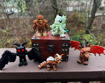 May include: A collection of six small, hand-painted figurines, including a brown yeti, a green monster, a black bat, a brown dragon, a brown and white dog, and a red dragon. The figurines are arranged on a wooden chest with a brass latch.