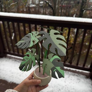May include: A green and white papercraft plant with a white pot. The plant is made of paper and has a realistic look. The plant is sitting on a wooden surface with snow on it.