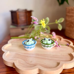 May include: Two small, light green frog figurines sit in miniature teacups on a wooden tray. One teacup has a blue rim and floral design, the other has a green rim and leaf design. The tray is shaped like a cloud. Flowers and greenery are in the background.