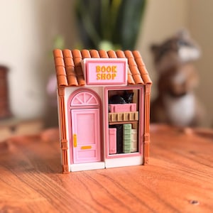 May include: A miniature pink and white book shop with a brown tile roof. The sign reads "BOOK SHOP". The shop has a pink door and a display window with books and a black cat. The shop is sitting on a wooden surface.