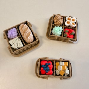 Puede incluir: Tres cestas de mimbre marrón en miniatura que contienen alimentos. Una cesta contiene una hogaza de pan, uvas moradas y trozos de queso. Otra contiene un cruasán, huevos duros, uvas verdes y fresas. La tercera contiene bayas y cubos de queso.