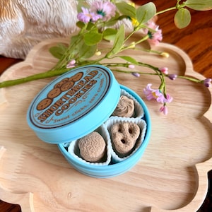 Puede incluir: Una lata azul claro de galletas de mantequilla con el texto "Shortbread Cookies" y "Sweet clicks, zero crumbs". La lata está abierta, revelando cuatro galletas de diferentes formas. La lata está sobre una bandeja de madera con flores.