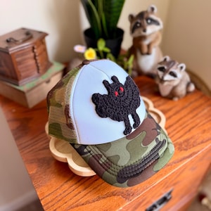 May include: A trucker hat featuring a white front panel with a black crocheted bat applique that has two small red eyes. The hat includes a green and brown camouflage pattern on the brim and side mesh panels, with a brown button on the top crown.
