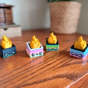 May include: Four small, colorful, square-shaped figurines with a flame design. Each figurine has a different color scheme, including black, pink, green, and blue, with a yellow flame on top. The figurines have cute, cartoon-like faces.