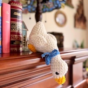 May include: A white crocheted goose plush toy with a textured yarn finish, featuring a bright yellow beak and feet, and a small blue ribbon tied around its neck. The goose has black plastic eyes and is positioned facing downward.