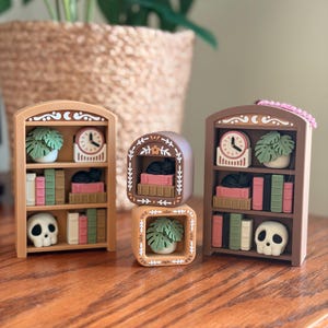 May include: Miniature wooden bookshelves with decorative elements. Each shelf features a clock, a potted plant, books, a skull, and a black cat. The shelves are in shades of brown, pink, green, and white, creating a whimsical aesthetic.