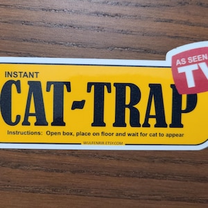 May include: A yellow and black sticker with the words "INSTANT CAT-TRAP" in bold letters. The sticker includes the text "Instructions: Open box, place on floor and wait for cat to appear." A red and white "AS SEEN ON TV" logo is in the upper right corner.