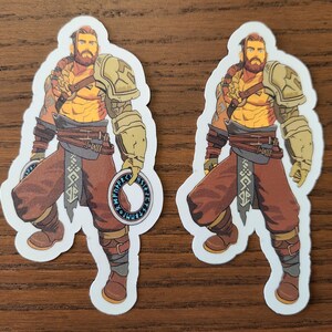 May include: Two stickers featuring a muscular warrior with a beard, wearing brown pants, armor, and holding circular objects. The warrior has tattoos and is depicted in a stylized illustration. The stickers are on a wooden surface.