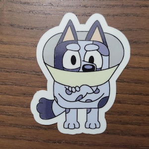May include: A sticker featuring a cartoon dog character with a light blue body, dark blue spots, and a light yellow cone. The dog has large black eyes and is smiling. The sticker is outlined in black.