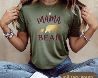 May include: A military green short-sleeve t-shirt featuring the text MAMA in a dark red script font, a gold silhouette of a large bear and a small cub, and the word BEAR in dark red block letters centered on the chest of the garment.