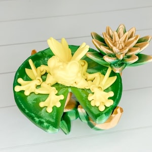 May include: A yellow frog figurine sitting on a green lily pad with a brown lily flower on top. The frog is posed with its legs outstretched and its mouth open.
