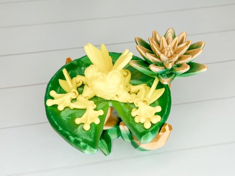 May include: A yellow frog figurine sitting on a green lily pad with a brown lily flower on top. The frog is posed with its legs outstretched and its mouth open.