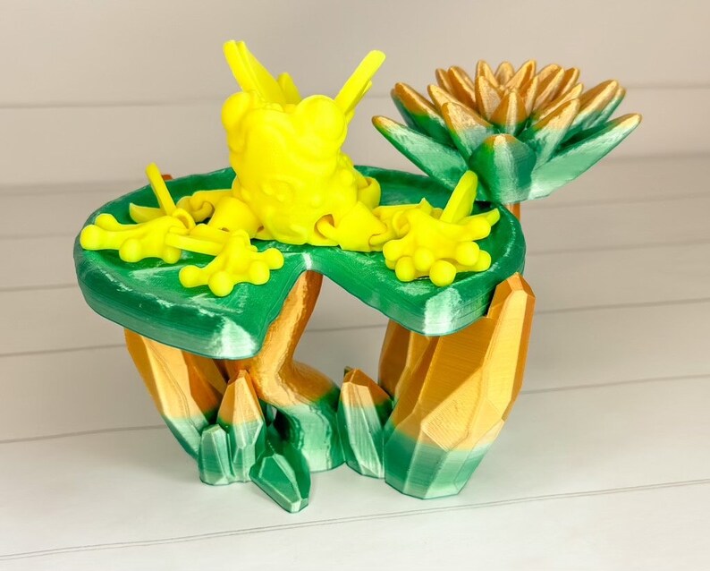 May include: A 3D printed sculpture of a frog sitting on a green lily pad with a golden flower. The frog is yellow and has a textured surface. The lily pad is green and has a textured surface. The flower is gold and has a textured surface.