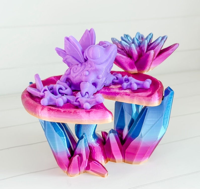 May include: A purple frog with wings sits on a pink and purple flower with blue and pink crystal accents.