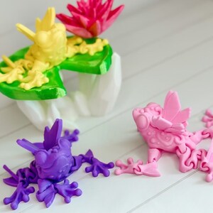 May include: Three 3D printed frogs, one yellow, one pink, and one purple, are posed on a white surface. The yellow frog is sitting on a green lily pad with a pink flower.