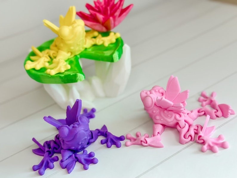 May include: Three 3D printed frogs, one yellow, one pink, and one purple, are posed on a white surface. The yellow frog is sitting on a green lily pad with a pink flower.
