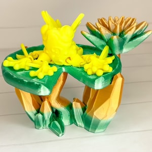 May include: A 3D printed sculpture of a frog sitting on a green lily pad with a golden flower. The frog is yellow and has a textured surface. The lily pad is green and has a textured surface. The flower is gold and has a textured surface.