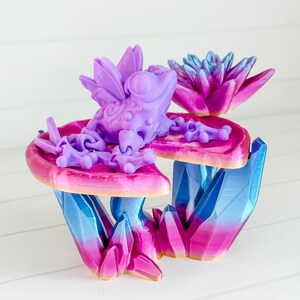 May include: A purple frog with wings sits on a pink and blue crystal base. The frog is perched on a pink flower-shaped platform.
