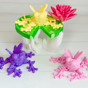 May include: Three 3D printed frogs, one yellow, one purple, and one pink. The yellow frog is sitting on a green lily pad with a pink flower. The frogs have articulated limbs and are posed in a playful manner.