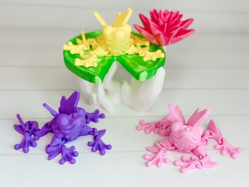 May include: Three 3D printed frogs, one yellow, one purple, and one pink. The yellow frog is sitting on a green lily pad with a pink flower. The frogs have articulated limbs and are posed in a playful manner.