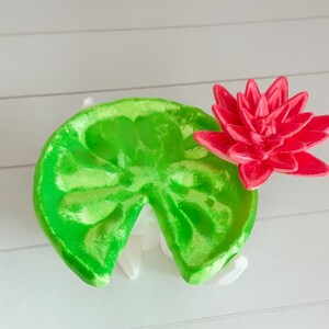 May include: A 3D printed green lily pad with a red lily flower on top. The lily pad is split in half and the flower is facing the right side of the image.