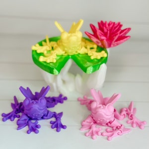 May include: Three 3D printed frogs, one yellow, one purple, and one pink, sitting on a white surface. The yellow frog is on a green lily pad with a pink flower.
