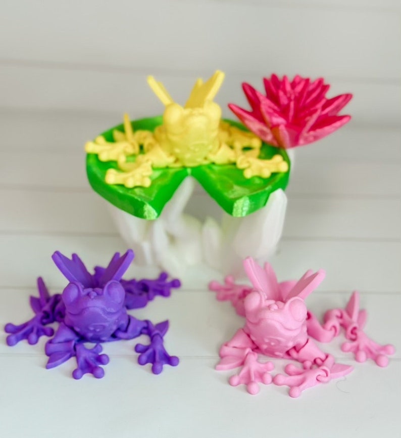 May include: Three 3D printed frogs, one yellow, one purple, and one pink, sitting on a white surface. The yellow frog is on a green lily pad with a pink flower.