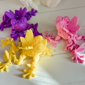 May include: Three articulated frog figurines, each with wings. The frogs are made of plastic and are painted in different colors: purple, pink, and yellow.