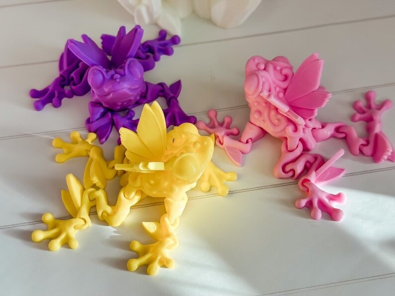 May include: Three articulated frog figurines, each with wings. The frogs are made of plastic and are painted in different colors: purple, pink, and yellow.