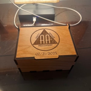 Peut inclure: Une boîte en bois avec une bordure noire et un motif gravé au laser. Le motif représente un triangle avec les mots "Unité", "Service" et "Rétablissement" à chaque point. À l'intérieur du triangle se trouve l'abréviation "AA" et la date "07/21/2023".