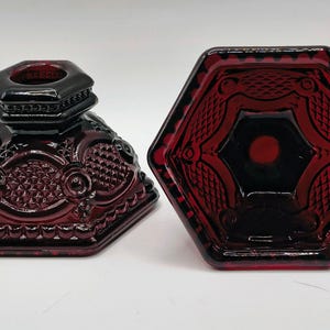 May include: Two deep red glass candle holders with a hexagonal base and a textured, pressed pattern. One holder is shown from a side angle, revealing a raised neck, while the other is viewed from directly above, displaying the geometric design.