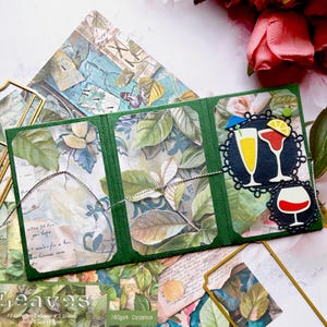 May include: A handmade green card with three clear pockets, filled with botanical-themed paper and leaves. The card is decorated with cocktail and wine glass cutouts. Additional botanical paper and a gold-colored frame are visible.