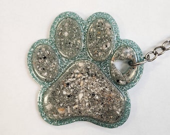 May include: A teal green glitter paw print keychain with a silver chain. The paw print is filled with a clear resin with embedded sand and small shells.