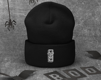 Könnte beinhalten: Eine schwarze Mütze mit einem weißen, gestickten Design am umgeschlagenen Rand. Das Wort "BOO" ist mit Schablonenbuchstaben geschrieben, und Fledermaus-Ausschnitte sind um die Mütze herum verstreut. Der Hintergrund hat ein strukturiertes, graues Aussehen, mit Spinnendekorationen.