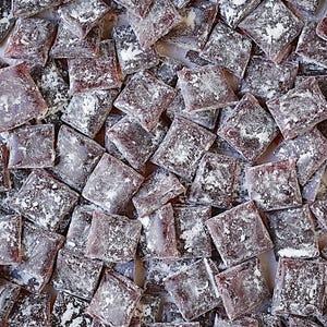 May include: Overhead shot of numerous square candies, each wrapped in clear packaging. The candies are a deep reddish-brown color and dusted with a generous coating of white powdered sugar, creating a visually appealing contrast. The candies are scattered.