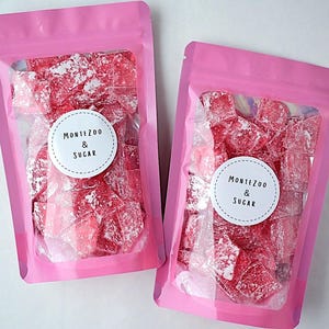 May include: Two pink pouches filled with red, sugar-coated candies. Each pouch has a white circular label with the text "MONTEZOO & SUGAR" in a simple font. The candies appear to be a type of rock candy.