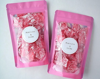 May include: Two pink pouches filled with red, sugar-coated candies. Each pouch has a white circular label with the text "MONTEZOO & SUGAR" in a simple font. The candies appear to be a type of rock candy.